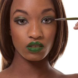 Young female beauty ethnic fashion model of African-American ethnicity with green lips and eye shades covered in dramatic make-up and holding makeup brush